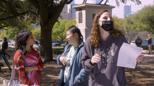 The author is a young woman in a grey sweatshirt with light brown curly hair. She is holding a piece of paper and wearing a black mask. She is standing on the Texas Capitol grounds with two other woman talking in the background.